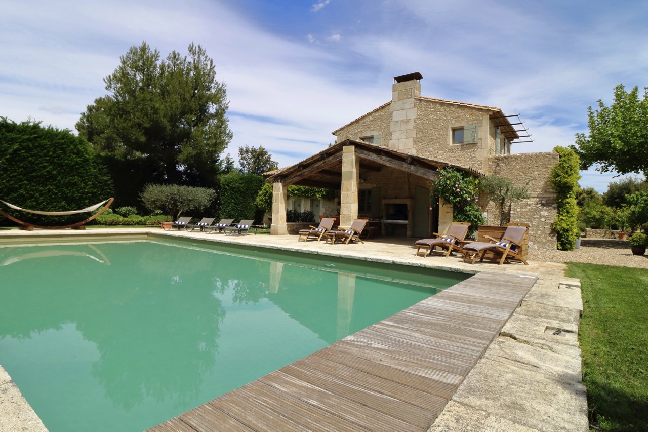 A luxurious stone house with a porch, next to a large swimming pool surrounded by loungers and a hammock, set in a lush garden under a clear blue sky.